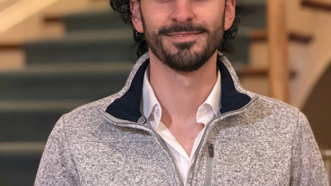 Portrait of Nikali Jordan wearing a light-gray quarter-zip sweater over a white collared shirt, standing indoors in front of a staircase. at Stagmere Manor